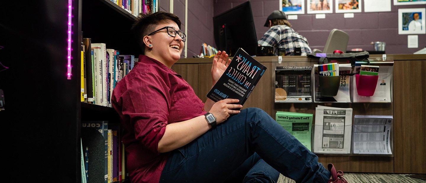 young adult seated in front of a book shelf reading a book called "Trans in College"