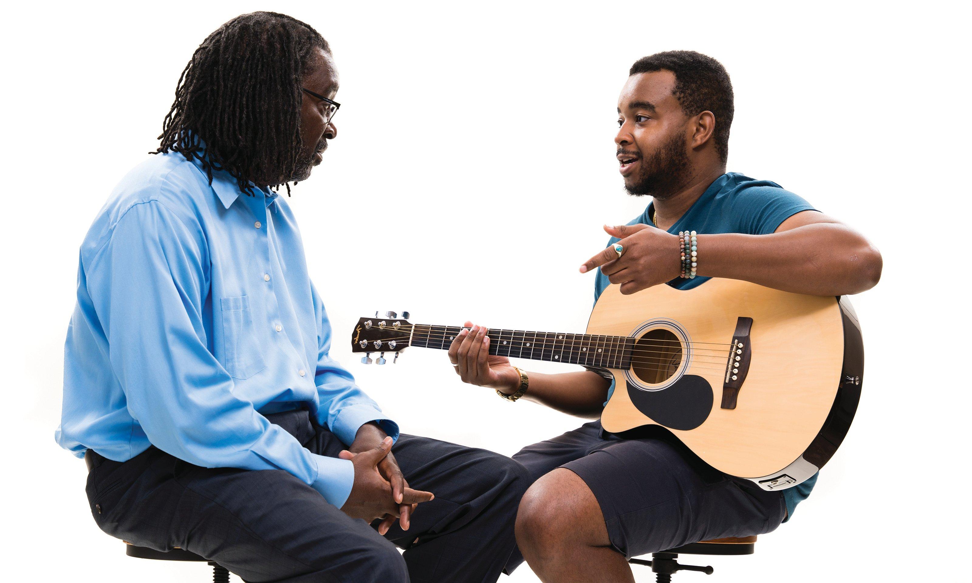 A man sitting and holding a guitar talking to another man.