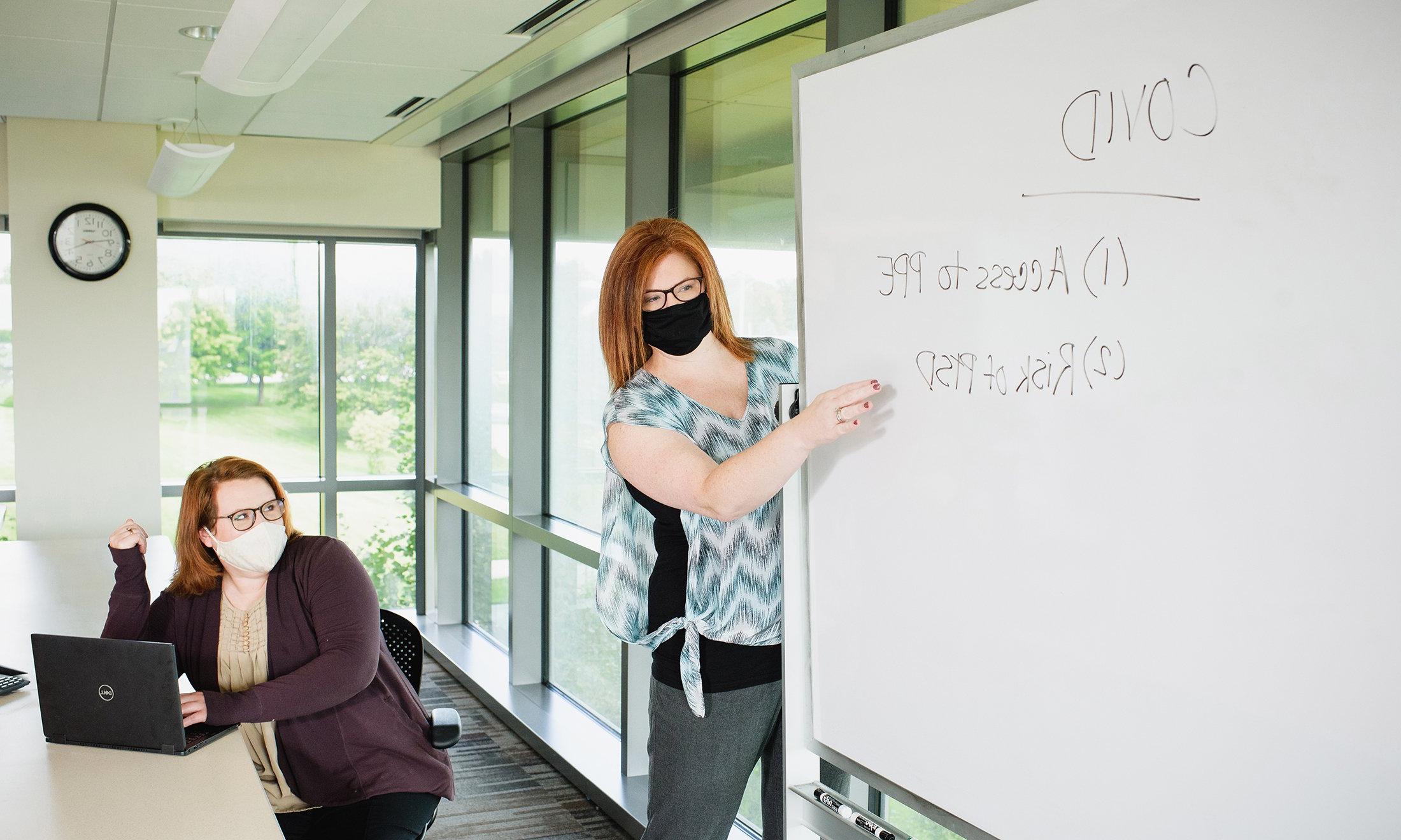 One woman writing on a white board, the other sitting. Both are wearing face masks.