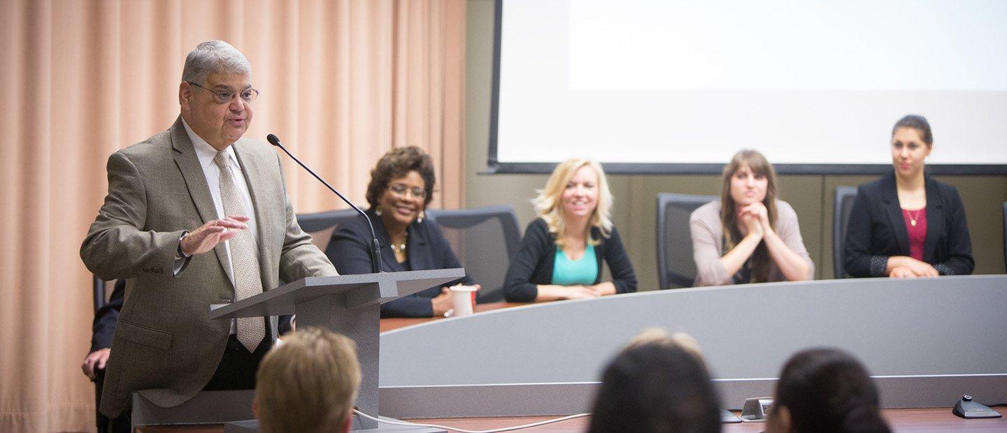 A man speaking into a microphone at a podium, with a panel of people seated behind him.